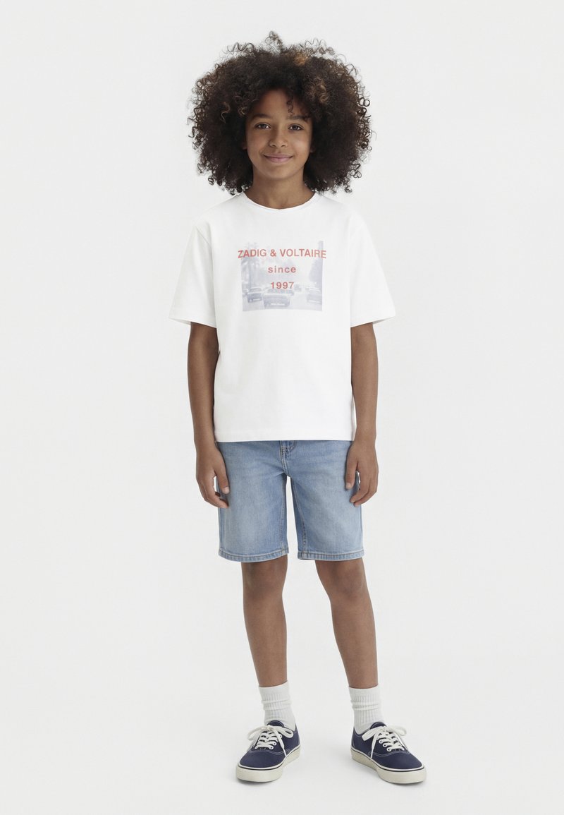 Dijete s kovrčavom kosom u bijeloj majici s grafičkim printom, svijetloplavim traper kratkim hlačama, bijelim čarapama i tamnoplavim tenisicama, stojeći na jednobojnoj pozadini.