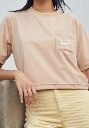 Person iført en beige Nike T-shirt med brystlomme og lysegule kordbukser, hvilende armen på en betonoverflade.