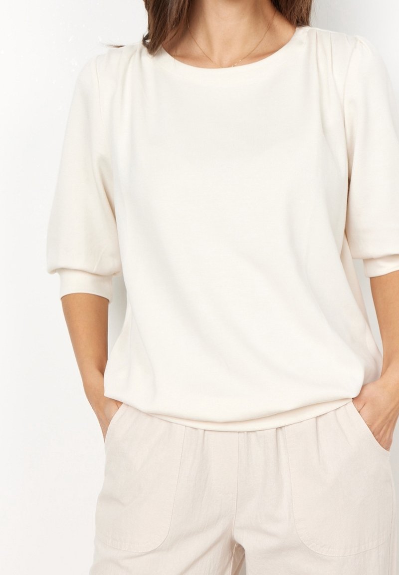 Mujer vestida con una blusa blanca de mangas abullonadas y pantalones beige claros, con las manos en los bolsillos, vista de los hombros a la cintura.