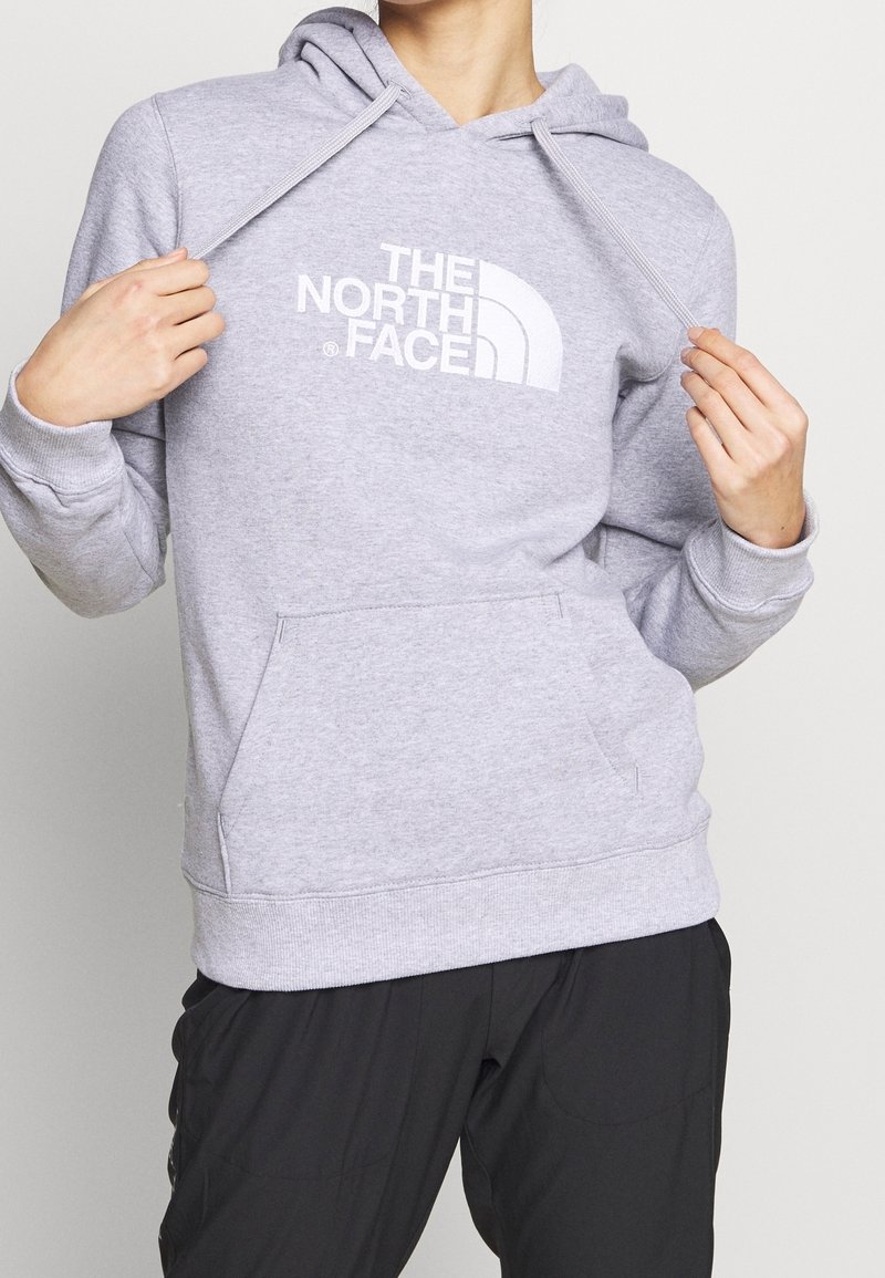 Personne portant un sweat à capuche gris clair The North Face tenant ses cordons, associé à un pantalon noir ample.