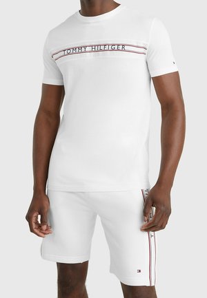 Mężczyzna w białym t-shircie Tommy Hilfiger i pasujących krótkich spodenkach z nazwą marki oraz paskami w kolorach czerwonym, białym i granatowym.