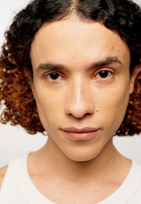 Närbild av en person med lockigt, axellångt hår som övergår från mörk till ljusbrun. Slät hy med subtil struktur.