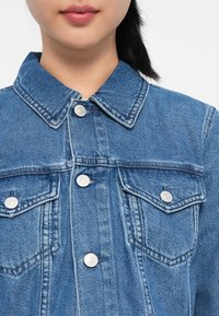 Blå denimjakke med klassisk krage, sølvknapper og to brystlommer. Har synlige sømmer og en lett falmet tekstur.