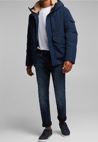 Mörkblå puffjacka med en mjuk beige innerväv, framfickor och knäppning med knappar. Kombinerad med mörka jeans och avslappnade skor.