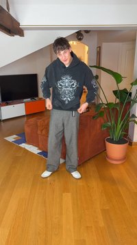 Camisola com capuz preta com design gráfico, calças cinzentas de perna larga, sapatilhas brancas, em pé numa sala com pavimento em madeira e uma planta verde.