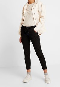 Ljusbeige denimjacka, tan t-shirt, svarta byxor med dragsko och vita sneakers med grå strumpor. Enkelt och figurnära design.