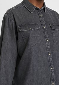 Camicia di jeans grigia scura con maniche lunghe, chiusura con bottoni e due tasche sul petto. Presenta un colletto classico e una texture morbida.