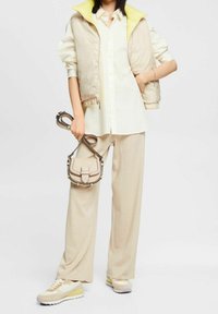 Gilet imbottito beige con fodera gialla, camicia bianca, pantaloni a gamba larga crema, sneakers beige con dettagli pastello, piccola borsa a tracolla.