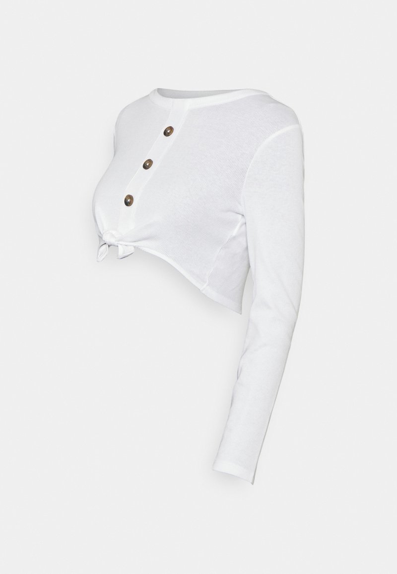Vit långärmad croppad topp med tre bruna knappar framtill och en knutdetalj vid nederkanten.