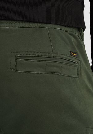 Los pantalones de algodón verdes presentan un bolsillo en la parte posterior con una solapa horizontal y una pequeña etiqueta de marca. La tela tiene una textura suave y mate.