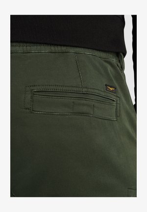 Los pantalones de algodón verdes presentan un bolsillo en la parte posterior con una solapa horizontal y una pequeña etiqueta de marca. La tela tiene una textura suave y mate.