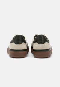 Chaussures en daim avec un dessus crème et des accents en cuir noir, de forme arrondie, et une semelle en caoutchouc brun. La vue de dos met en valeur des détails texturés.