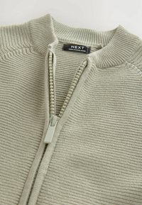 Hellgrüner Reißverschluss-Pullover mit gerippter Textur, ausgestattet mit einem hohen Kragen und einem Kunststoff-Reißverschluss, der entlang des Schiebers mit Perlen verziert ist. Markenetikett sichtbar.