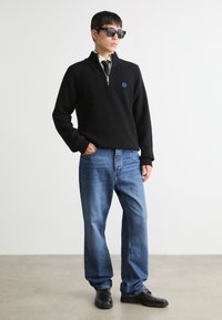 Maglione a zip nera in maglia con texture a coste, logo blu, jeans di denim blu e scarpe nere, completato da occhiali da sole neri.