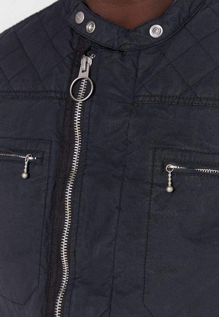 Schwarze gesteppten Jacke mit hohem Kragen, silbernen Reißverschlüssen und Druckknöpfen. Verfügt über zwei zipbare Taschen mit Metallgriffen.
