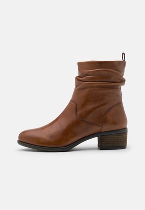 Botte bottine en cuir marron avec talon bas empilé, tige souple et languette à l'arrière sur un fond clair uni.