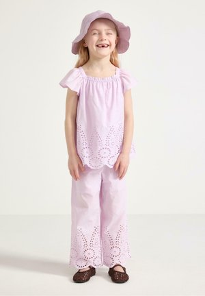 Hymyilevä nuori tyttö, jolla on päälleen sopiva vaaleanvioletti, reikäkangasyläosa ja housut, aurinkohattu sekä tummat punotut kengät, seisoo yksinkertaisen taustan edessä.