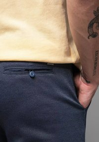 Pantaloni navy a trama con chiusura a bottone nella tasca posteriore, abbinati a una T-shirt di cotone giallo chiaro. Dettaglio in primo piano.