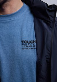 Blå bomullst-shirt med svart text "TOUGH TRAILS" på bröstet, stylad under en mörk jacka. Mjukt tyg, rund halsringning.