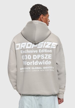 Tânăr cu păr creț poartă hanorac bej cu text alb "DROPSIZE Exclusive Edition 030 DPSZE Worldwide" pe spate, mâinile în buzunare.