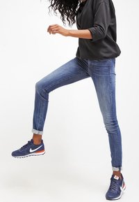 Vaqueros skinny de mezclilla azul con un diseño clásico de cinco bolsillos, con los puños enrollados, combinados con zapatillas deportivas oscuras que tienen detalles en blanco y rojo.