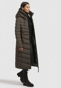 Giacca a vento lunga color verde oliva con colletto alto, motivo trapuntato, chiusura con zip davanti e tasche laterali, abbinata a stivali neri alla caviglia.