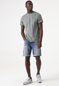 T-shirt verde chiaro con motivo e colletto a girocollo, abbinata a pantaloni corti in denim blu sfilacciati arrotolati in fondo e sneakers grigie.