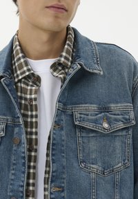 Džinsinis švarkas su šviesiai mėlynu skalbimu, turintis mygtukais užsegamus krūtinius kišenes; dėvimas virš tamsiai žalios ir juodos languotos flanelės marškinėlių.