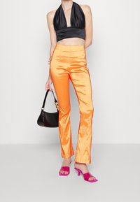 Svart halterneck crop top, glänsande orange satinbyxor, rosa klacksandaler och en svart texturerad handväska. Släta texturer och livfulla färger.