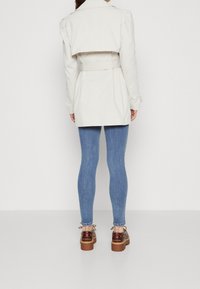 Trench ligero color beige con cintura ceñida, hombreras y cuello alto, combinado con jeans skinny azules y zapatos de plataforma marrones.