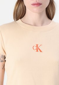 Beige geribbelde t-shirt met een ronde hals. Heeft een klein rood en wit "Calvin Klein" logo op de borst. Soepele textuur.