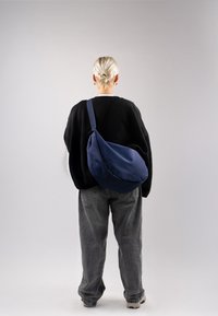 Borsa a spalla in tessuto navy con design curvo, indossata sopra un maglione nero oversize e pantaloni grigi larghi. Texture liscia, senza hardware visibile.