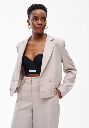 Kobieta z krótkimi włosami, ubrana w beżowe spodnie garniturowe i marynarkę, na czarnym krótkim topie z napisem "God save queens", pozująca z rękami w kieszeniach.