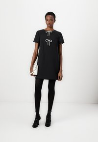 Abito nero a maniche corte con scollatura rotonda, caratterizzato da un dettaglio decorativo a fiocco sul davanti. Indossato con collant neri e scarpe con tacco.