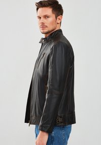 Chaqueta de cuero negro con cuello, cremallera frontal y bolsillo en el pecho. Presenta un acabado texturizado y detalle de hebilla en la parte inferior.