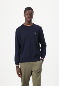 Marineblauwe pullovertrui met lange mouwen, geribde kraag met oranje, groene en rode strepen; Lacoste-logo geborduurd op de borst.