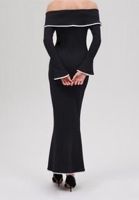 Vrouw in een aansluitende zwarte off-shoulder jurk met witte bies, belmouwen en zwarte hoge hakken, staande met haar handen gekruist achter haar rug.