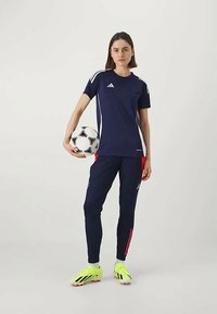 Noor naine tumesinistes Adidas spordiriietes hoiab jalgpalli, kandes erekollaseid satsisid, seistes ühtlases valges taustas.