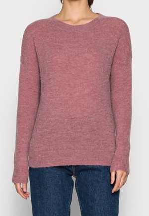 Femme portant un pull à col rond à manches longues de couleur rose pâle, associé à un jean en denim bleu foncé, posant devant un fond uni.