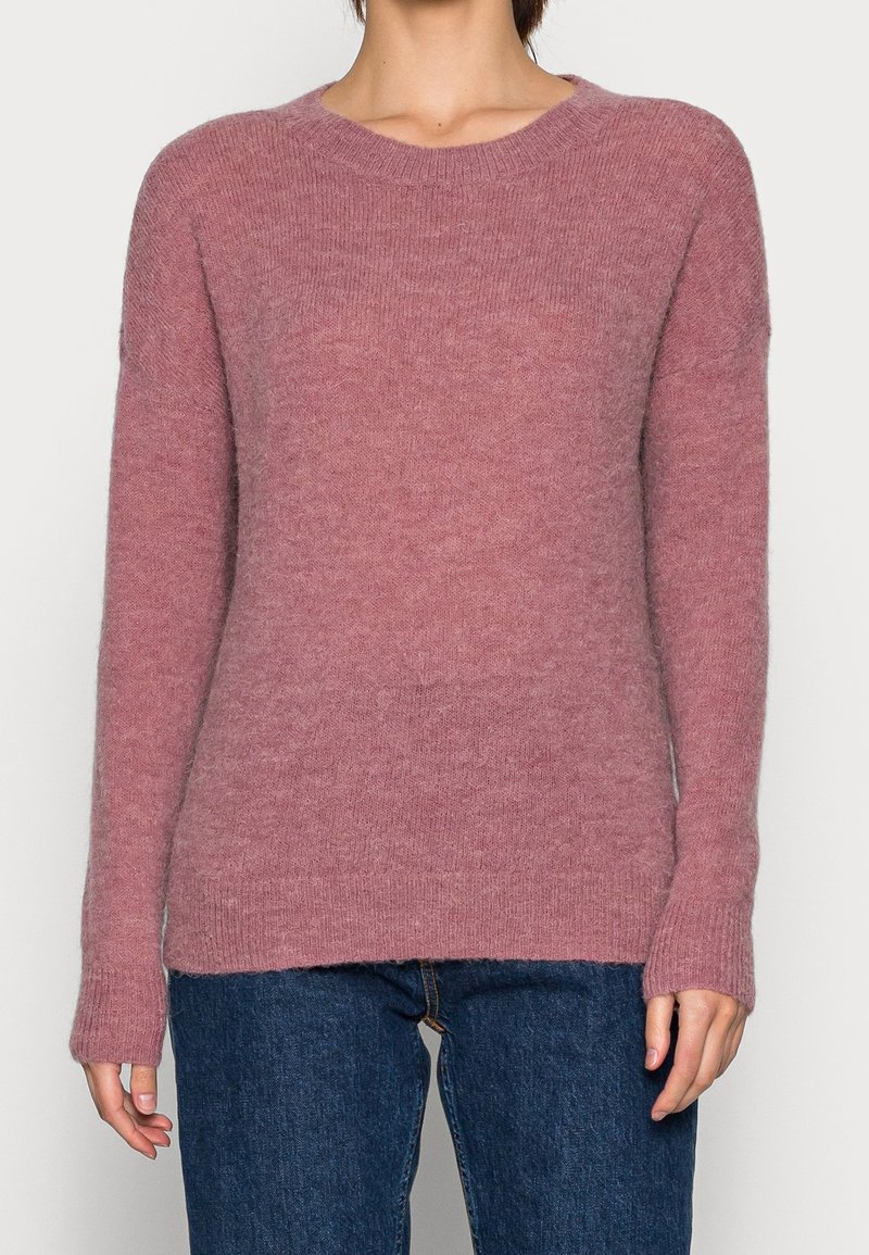 Femme portant un pull à col rond à manches longues de couleur rose pâle, associé à un jean en denim bleu foncé, posant devant un fond uni.