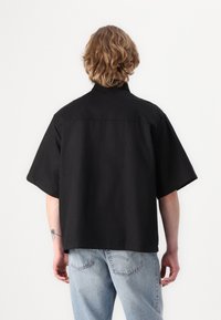 Camisa preta de manga curta com um corte descontraído, apresentando um colarinho elevado e uma textura suave. Combinada com jeans de ganga azul clara.