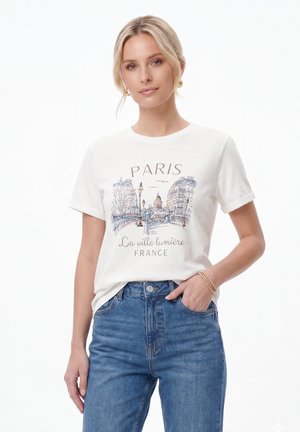 Femeie blondă purtând un tricou alb cu grafică tematică Paris și blugi albaștri, pozând cu o mână în buzunar și un zâmbet ușor.