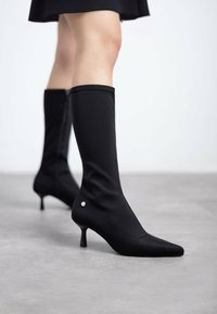 Botas negras hasta la rodilla con una punta ajustada y afilada, hechas de un material suave, que presentan un pequeño acento plateado cerca del tacón y un tacón de aguja.
