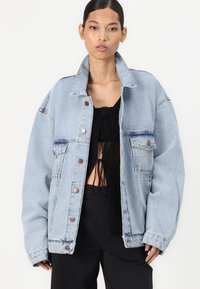 Svetlo modra oversize denim jakna s srebrnimi pritisnimi gumbi, žepoma na prsih in ovratnikom, ki je zasnovana s sproščenim krojem in rahlo raztrganim robom.