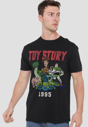 Ein Mann trägt ein schwarzes Toy Story 1995 T-Shirt mit Woody, Buzz Lightyear, Rex, Hamm und drei grünen Aliens auf grauem Hintergrund.