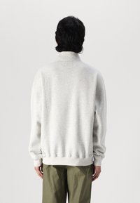 Sudadera gris claro con un collar alto, mangas largas y puños de canalé. Tejido suave con un corte relajado, llevada sobre pantalones verde oliva.