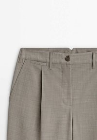 Pantalon gris coupé sur mesure avec passants de ceinture, fermeture à boutons sur le devant, poches latérales et pli frontal sur fond blanc.