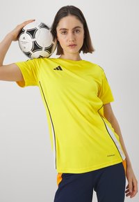 Geltona sportinė marškinėlė su juodais akcentais ir Adidas logotipu, trumpomis rankovėmis, minkšta medžiaga. Modelis laiko juodai baltą futbolo kamuolį.