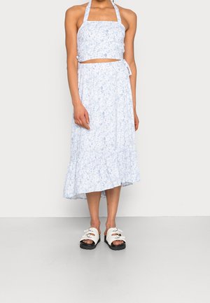 Mujer con top corto halter blanco y azul claro con estampado floral y falda midi a juego con bajo con volantes, combinados con sandalias blancas con hebilla.