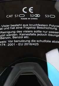 Czarna osłona z poliwęglanu z powłoką zapobiegającą parowaniu; zawiera certyfikaty bezpieczeństwa (CAT S1, S2) oraz instrukcje dotyczące pielęgnacji w języku niemieckim.